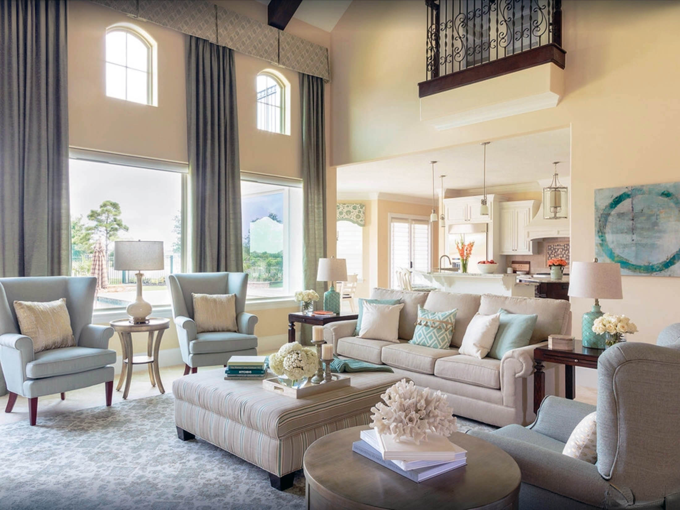 Elegant living room with light-colored sofas, armchairs, and decor, complemented by fresh interior painting. Large windows with curtains offer an open view to a stylish kitchen and dining area in the background.
