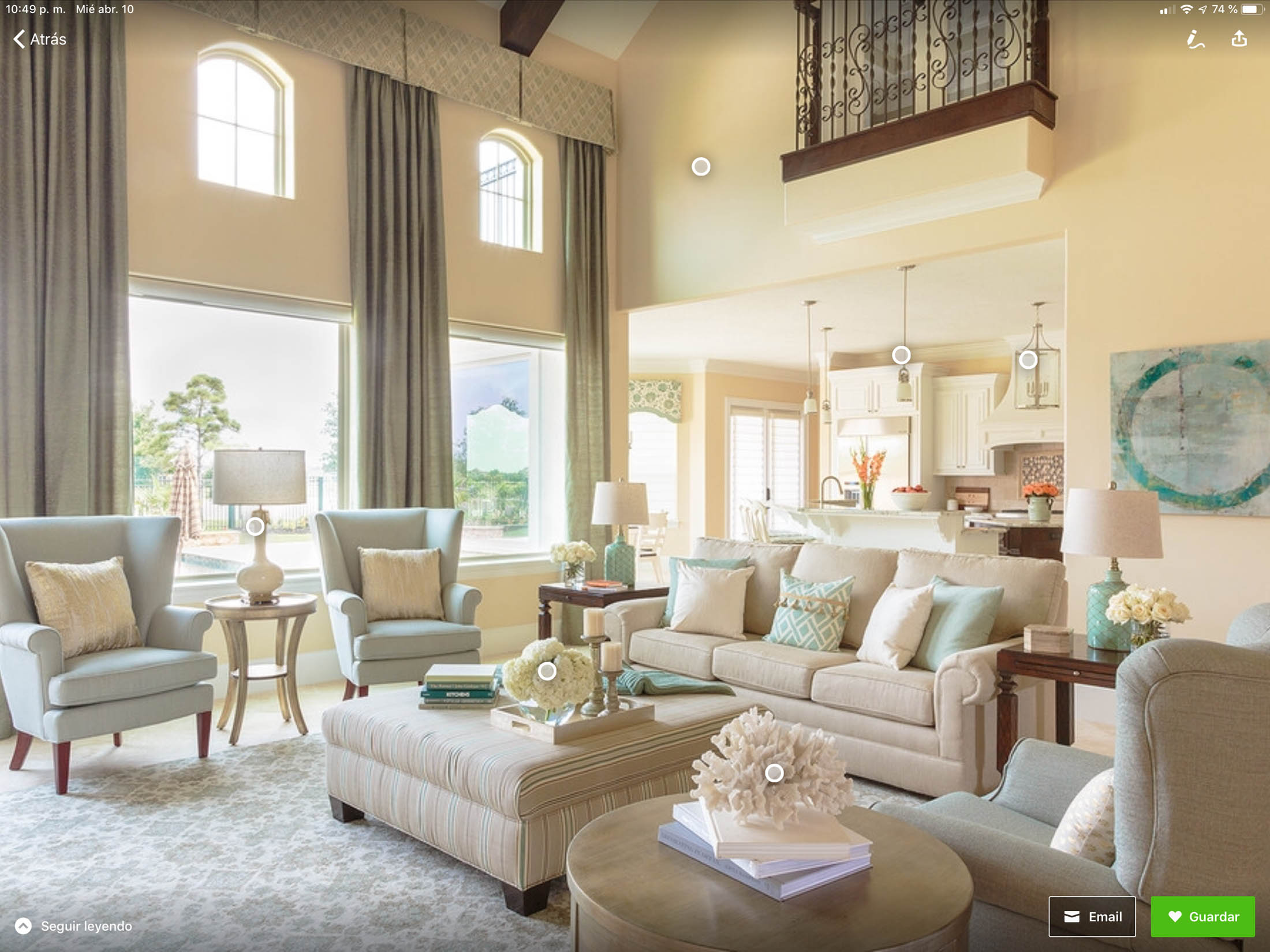 Spacious, well-lit living room with neutral-toned sofas, armchairs, large windows, a tufted ottoman, and a view into a white kitchen with bar stools and pendant lights.