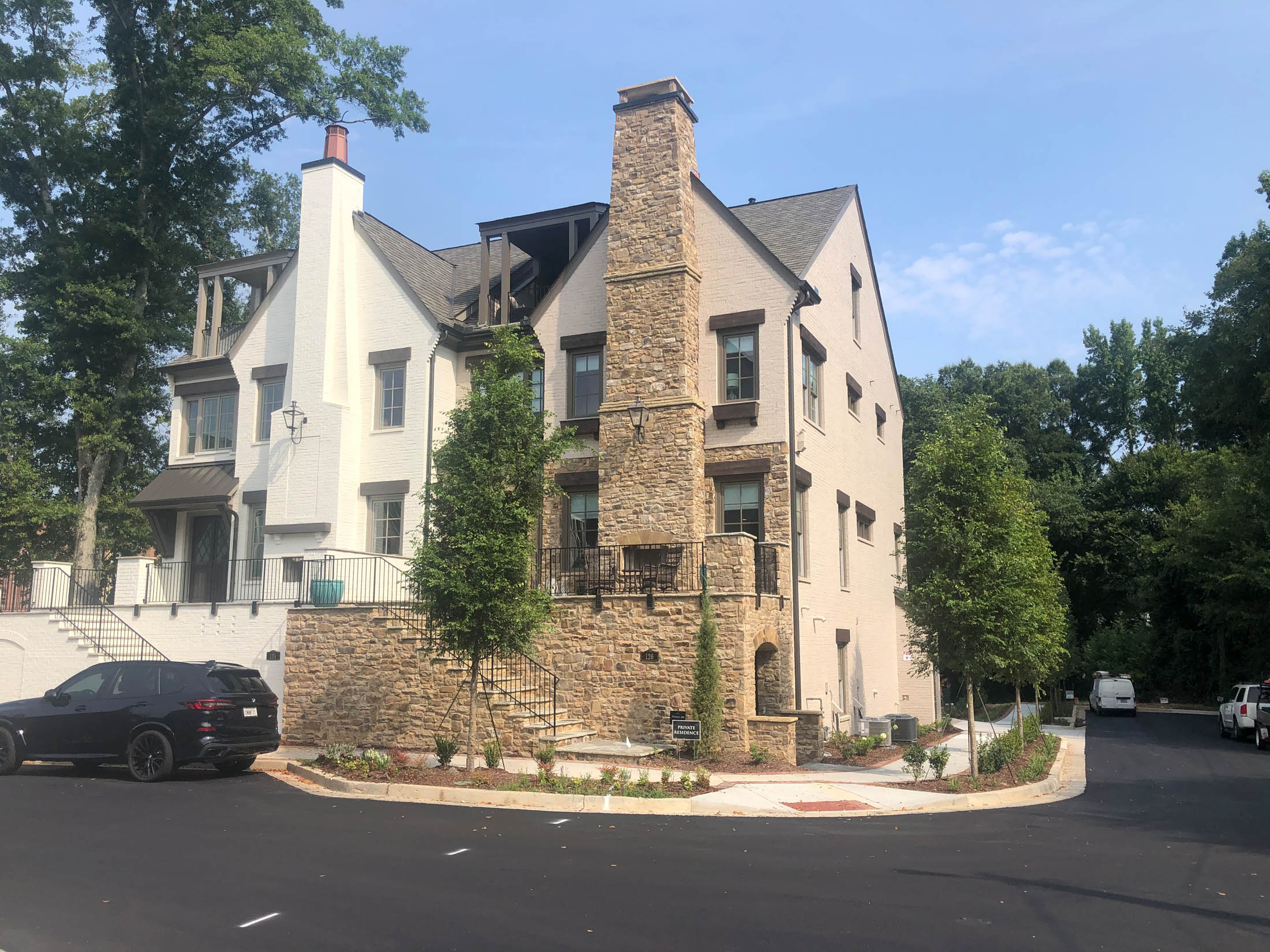 Three-story modern house with stone and white exterior, multiple balconies, and a tall chimney, situated at a corner on a newly paved residential street with parked vehicles.
