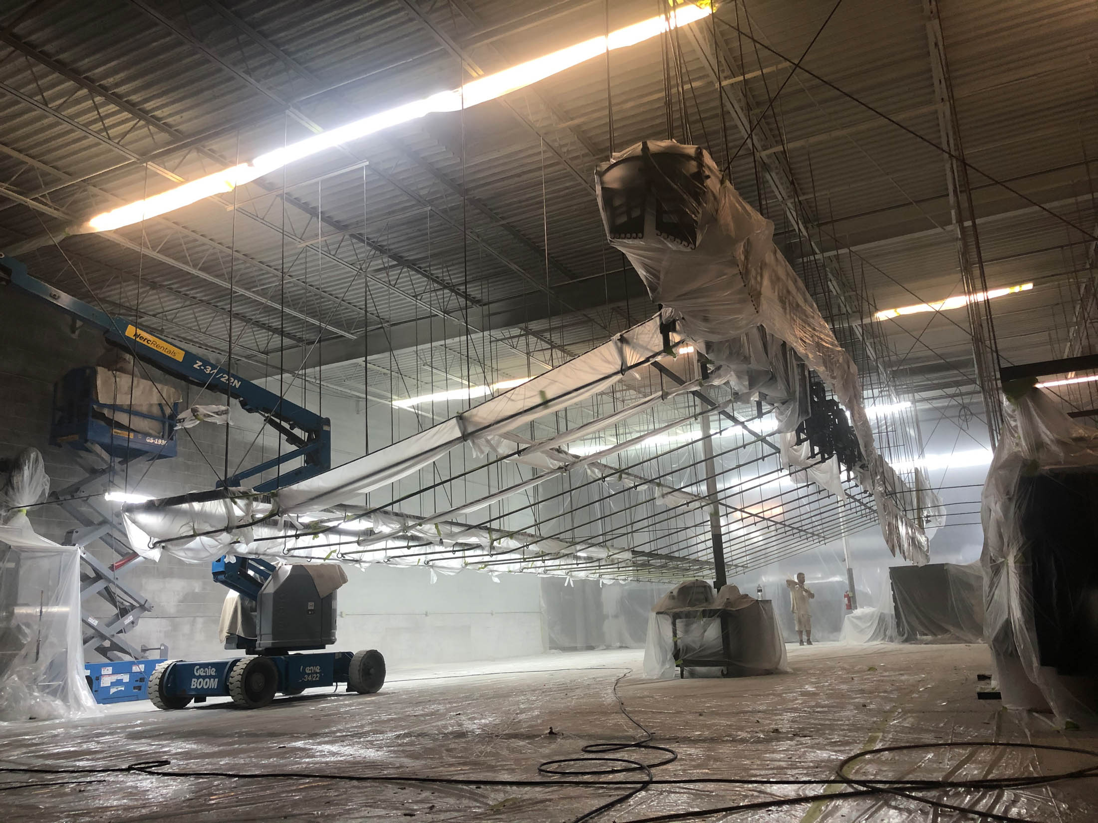 Large indoor construction site with overhead ducts wrapped in plastic, workers using lifts, and protective floor coverings throughout the space.