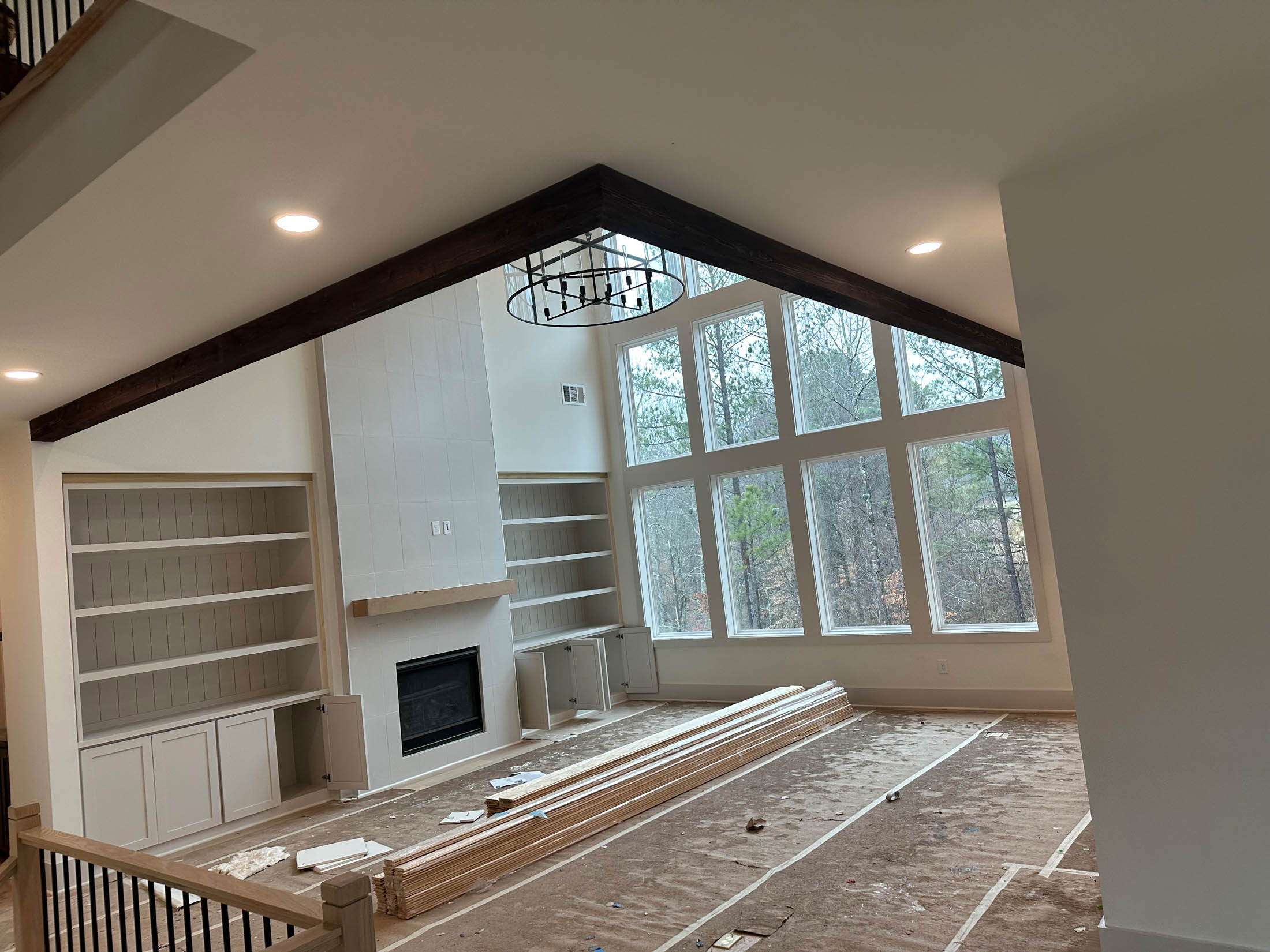 A spacious, unfinished living room with large windows, built-in shelves, a fireplace, exposed beam ceiling, and construction materials scattered on the floor.