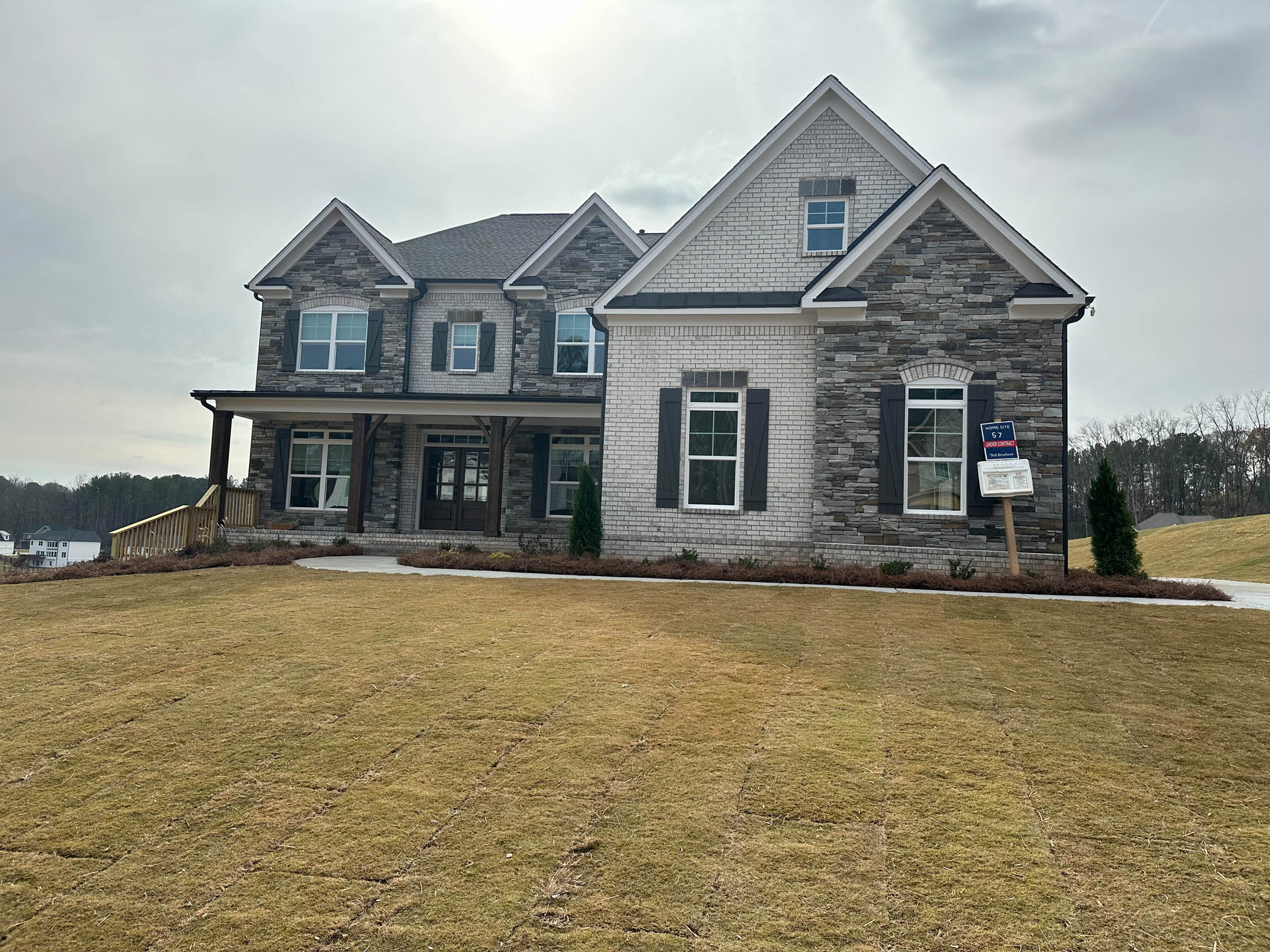 Two-story house with stone and brick exterior, large front yard, and a "For Sale" sign by the driveway under a cloudy sky.