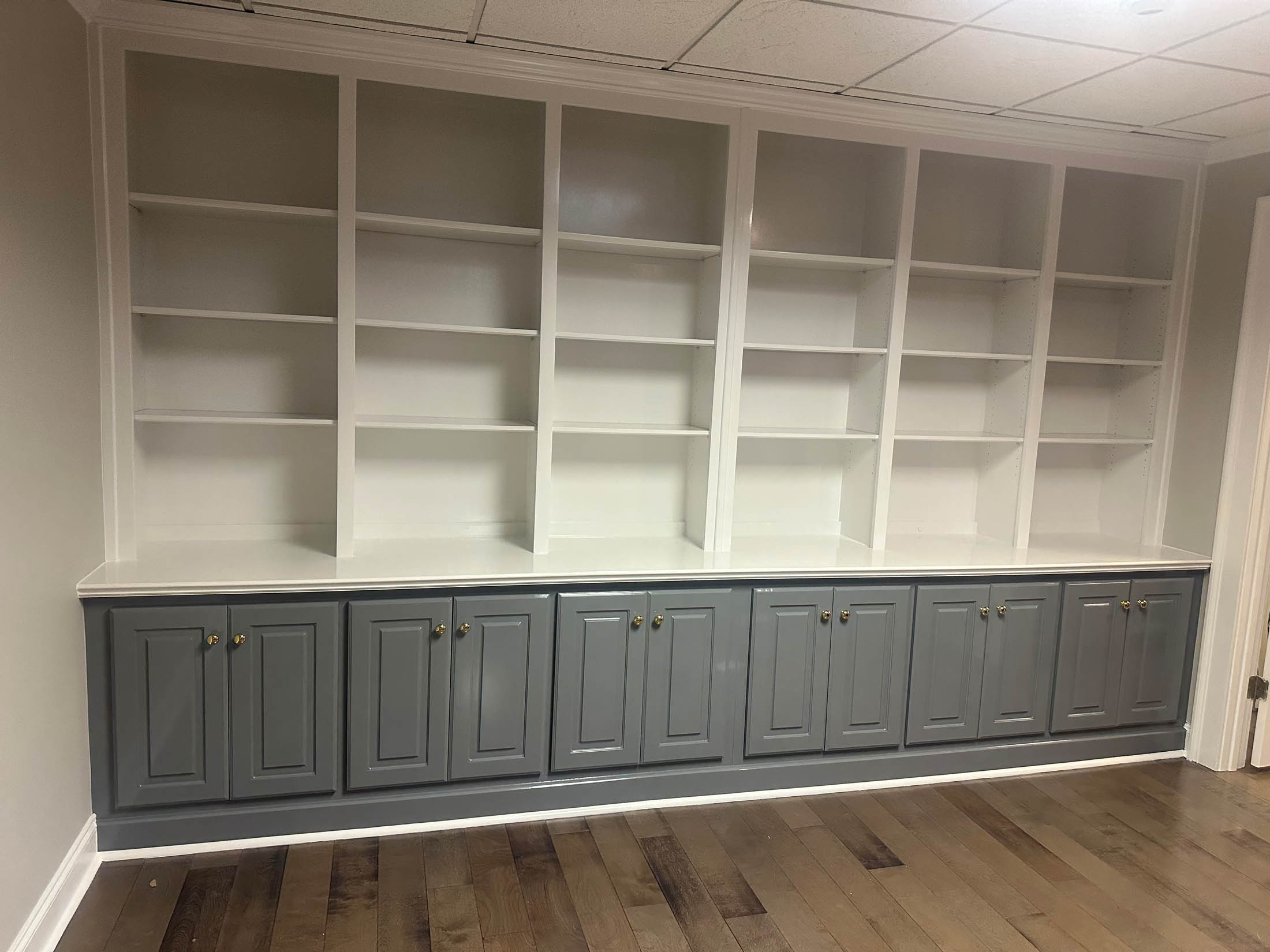 Built-in shelving unit with white open shelves above and gray cabinets with doors below, set against a beige wall and hardwood floor.