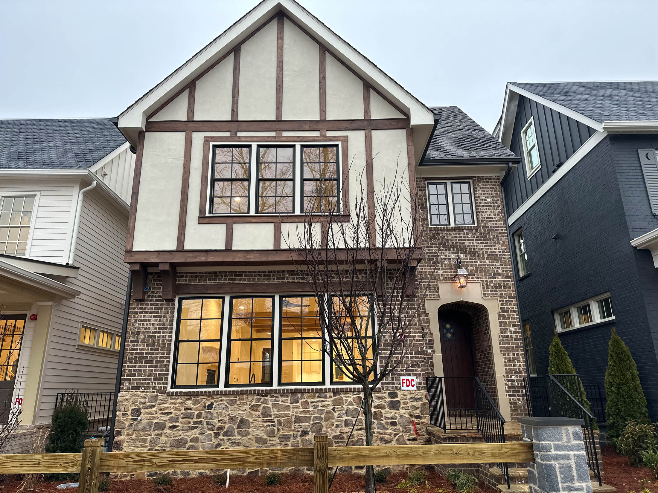 A two-story house with Tudor-style architecture, featuring brick and stone exterior, large front windows, and a leafless tree in the front yard.