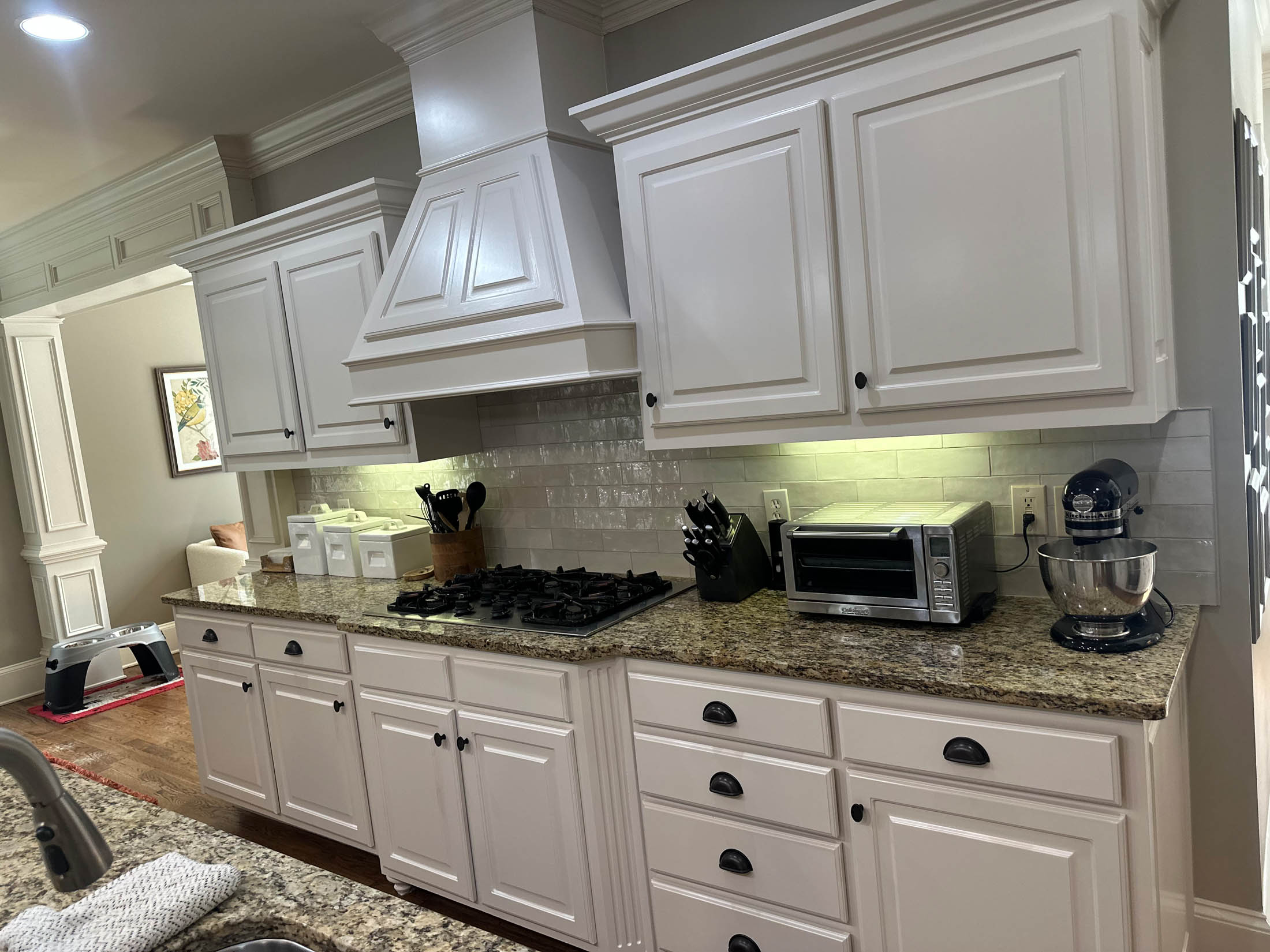 White kitchen cabinets with granite countertops, a gas cooktop, toaster oven, stand mixer, and utensil holders. Under-cabinet lighting highlights a tiled backsplash.