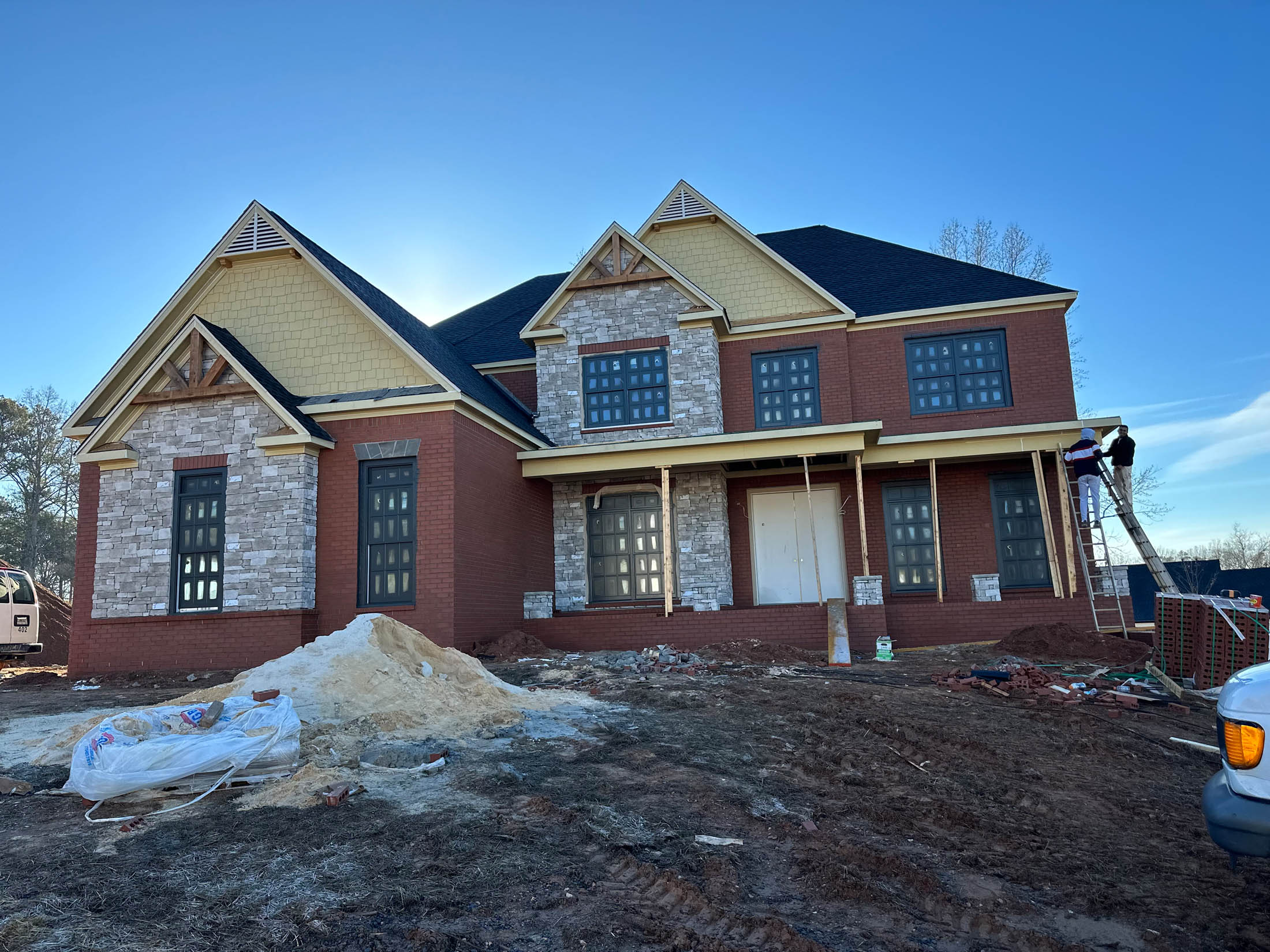 Two workers are on a ladder installing trim on the front porch of a partially completed brick house at a construction site.