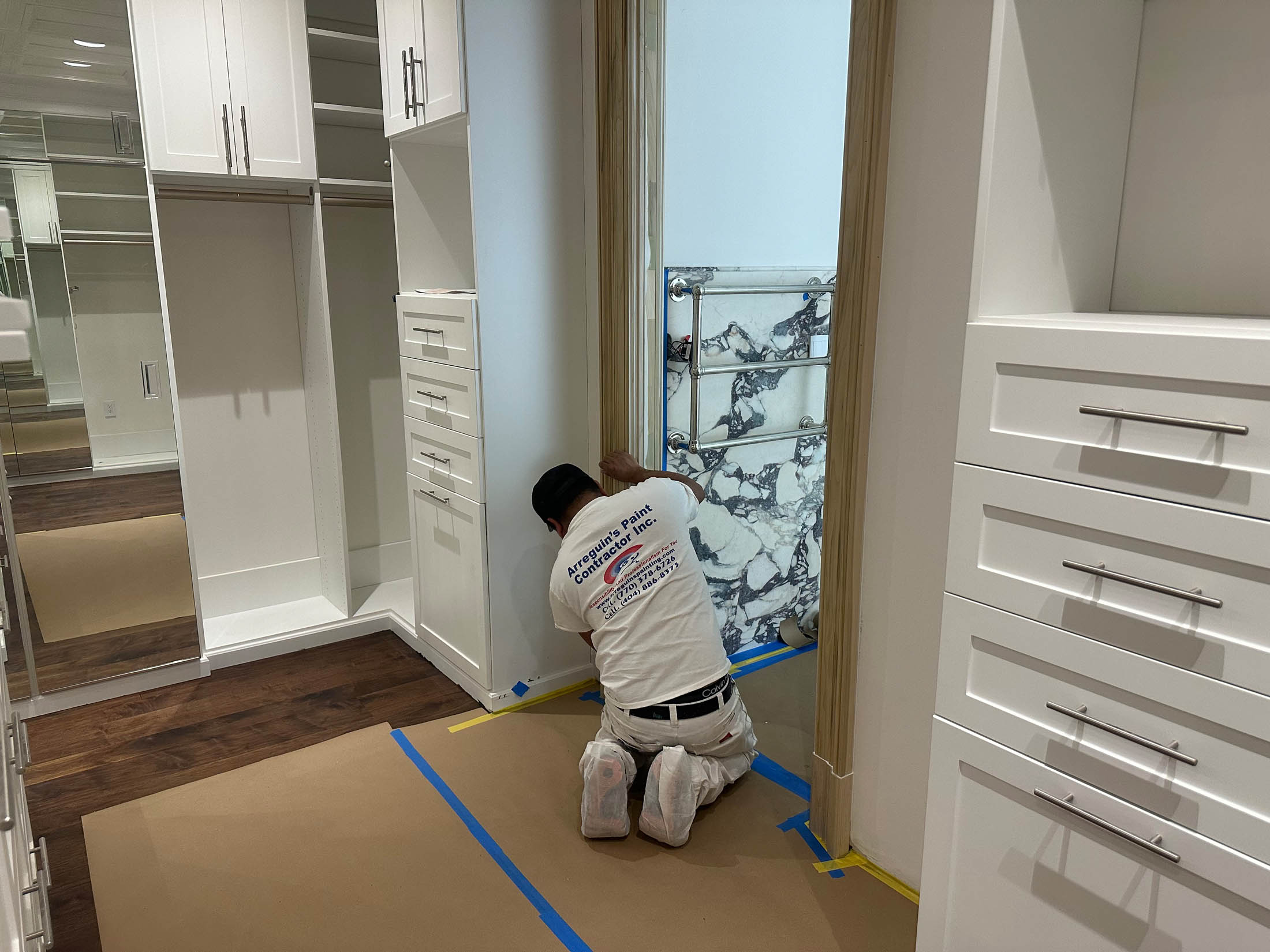 A worker from a painting company in Marietta kneels on the floor in a walk-in closet, preparing to paint or perform maintenance near a doorway, with cabinets and drawers visible.