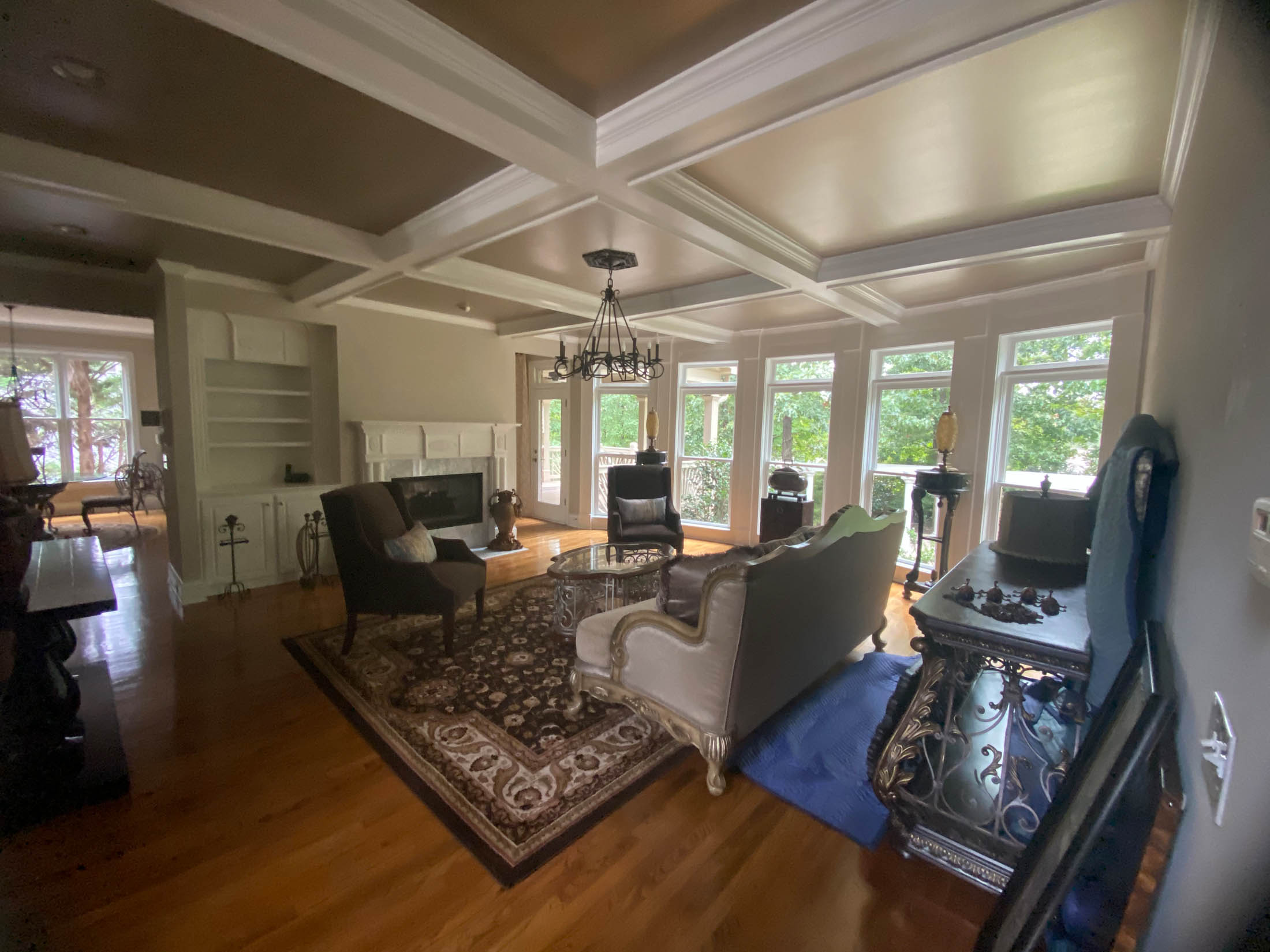 Spacious living room with hardwood floors, large windows, coffered ceiling, fireplace, and classic furniture including a sofa, armchairs, and a rug.