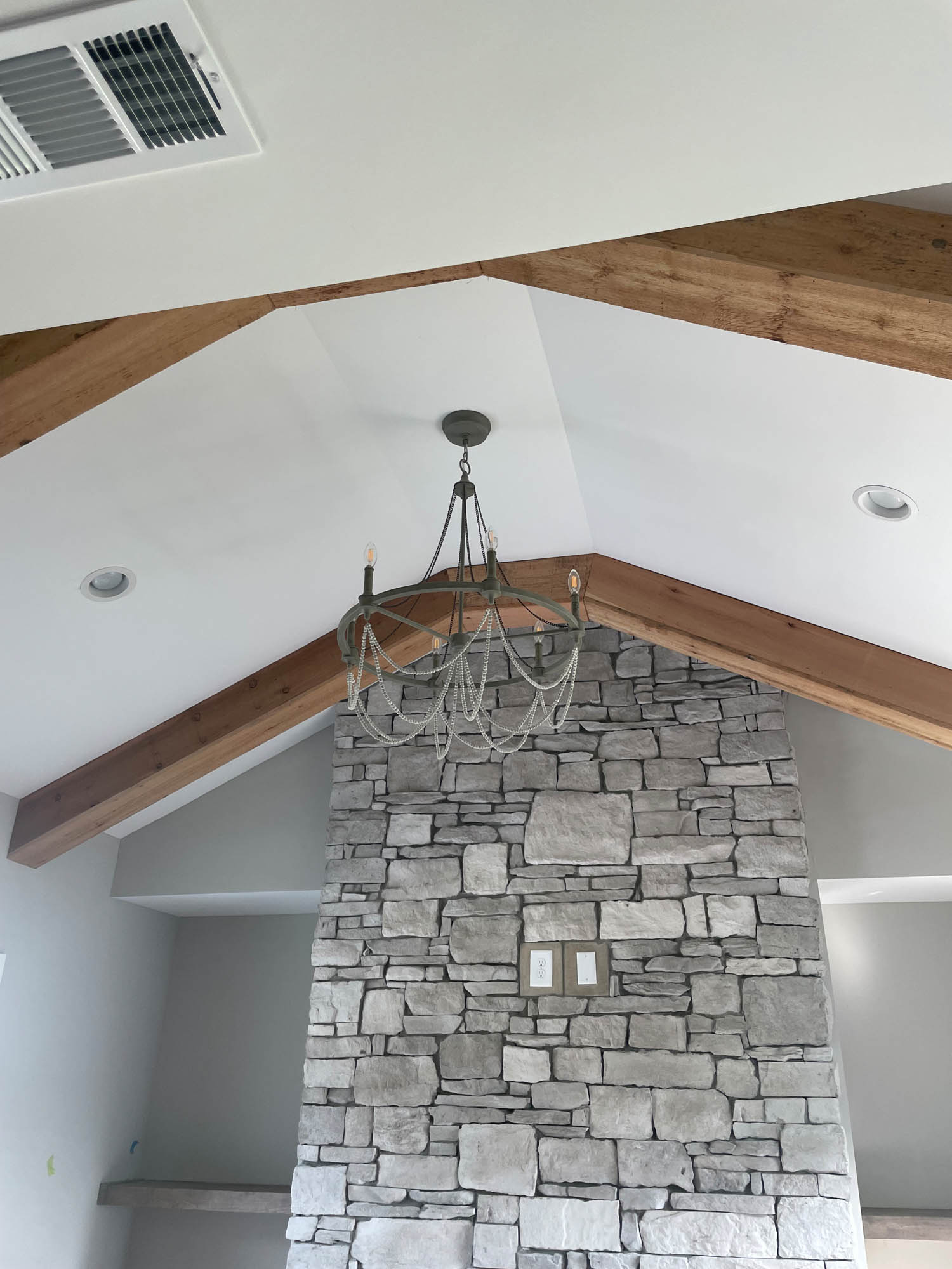 Ceiling with exposed wooden beams, a stone accent wall, a modern chandelier, and recessed lighting in a neutral-toned room.