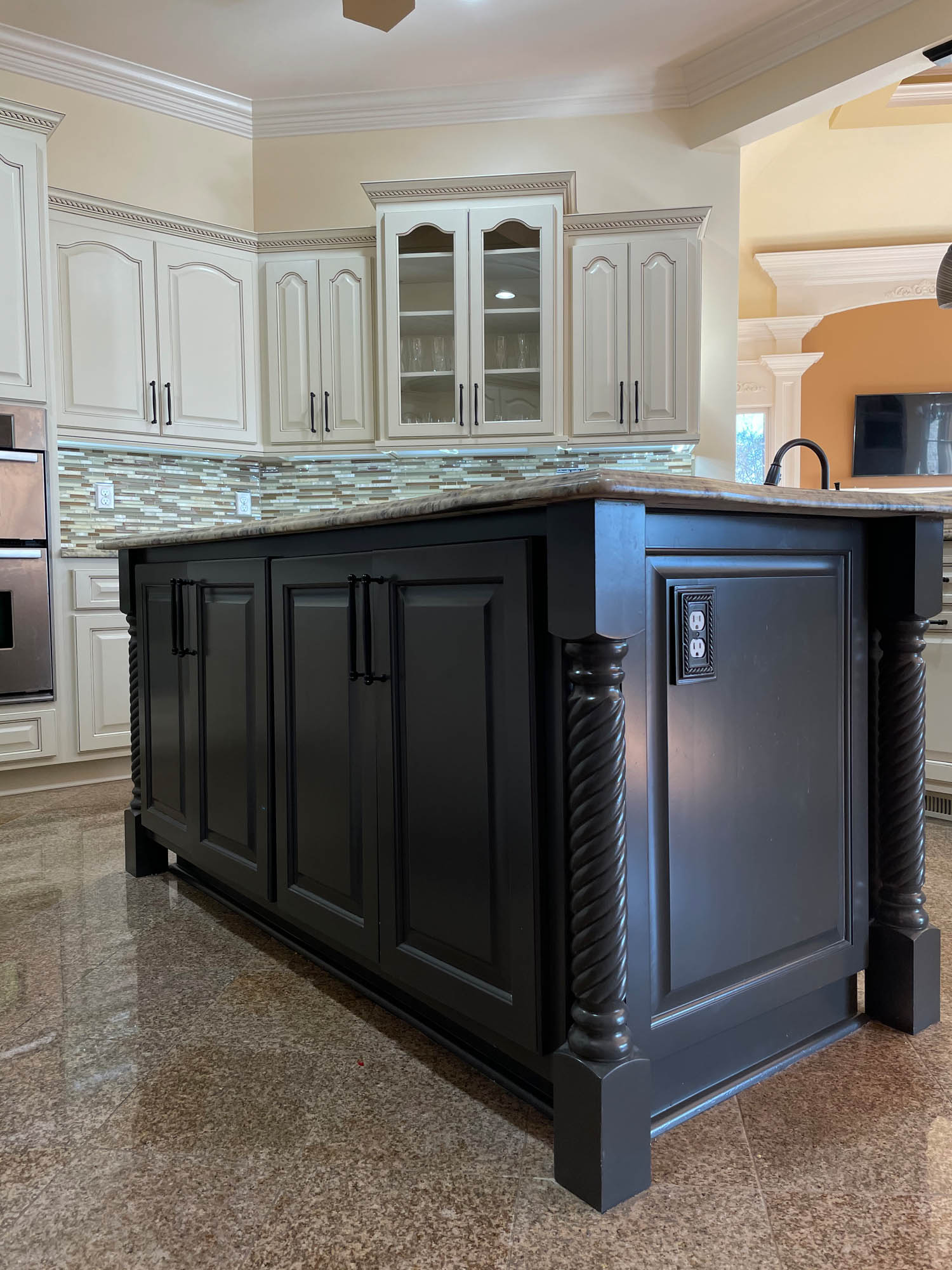 Kitchen with a large black island featuring decorative legs, white cabinetry, glass cabinet doors, a tile backsplash, and polished stone flooring.
