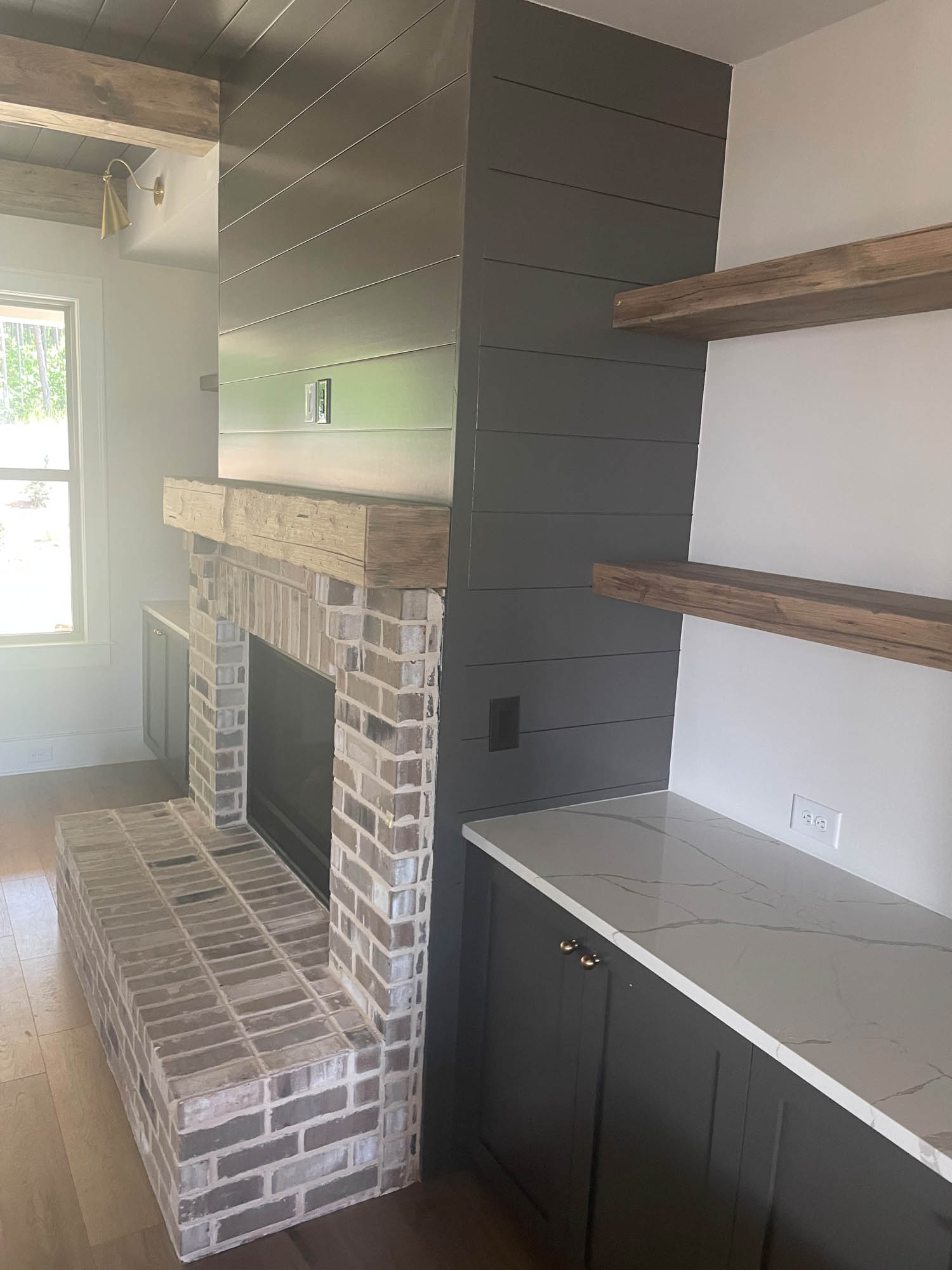 A brick fireplace with a wooden mantel, dark shiplap paneling, built-in shelves, and a countertop with cabinets in a modern living space.