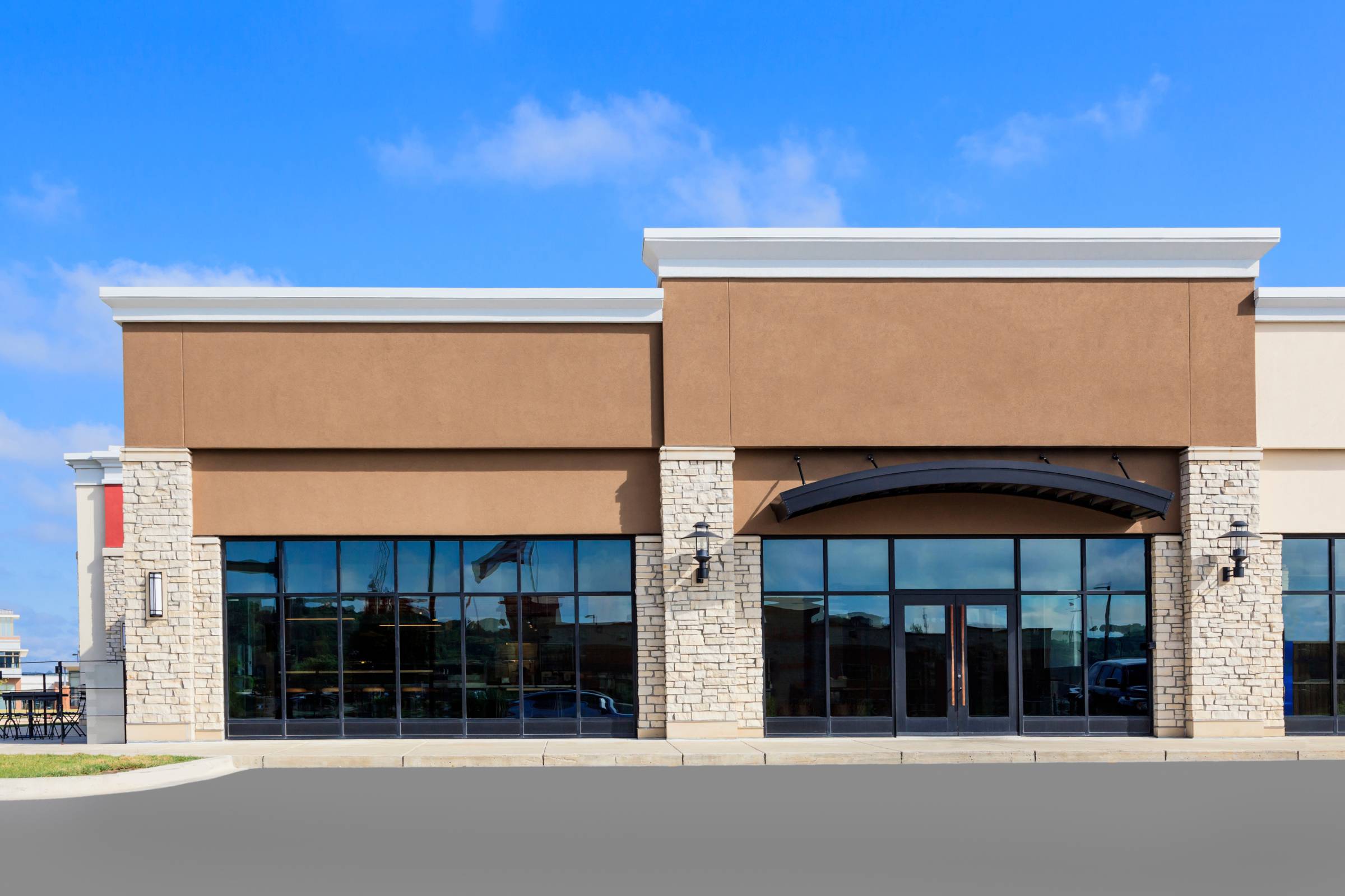 A modern, empty commercial storefront with large glass windows, beige and brown walls, stone accents, and a clear blue sky above—perfectly showcasing flawless new construction painting throughout the space.