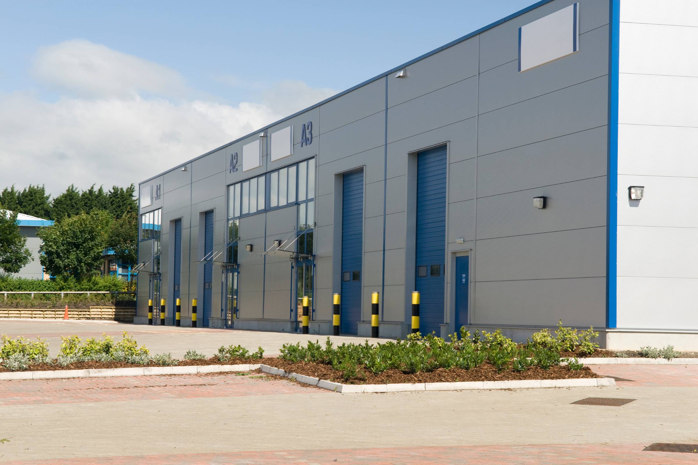 A modern industrial warehouse building with multiple loading bays, blue doors, yellow bollards, and landscaped areas in front featuring fresh new construction painting.