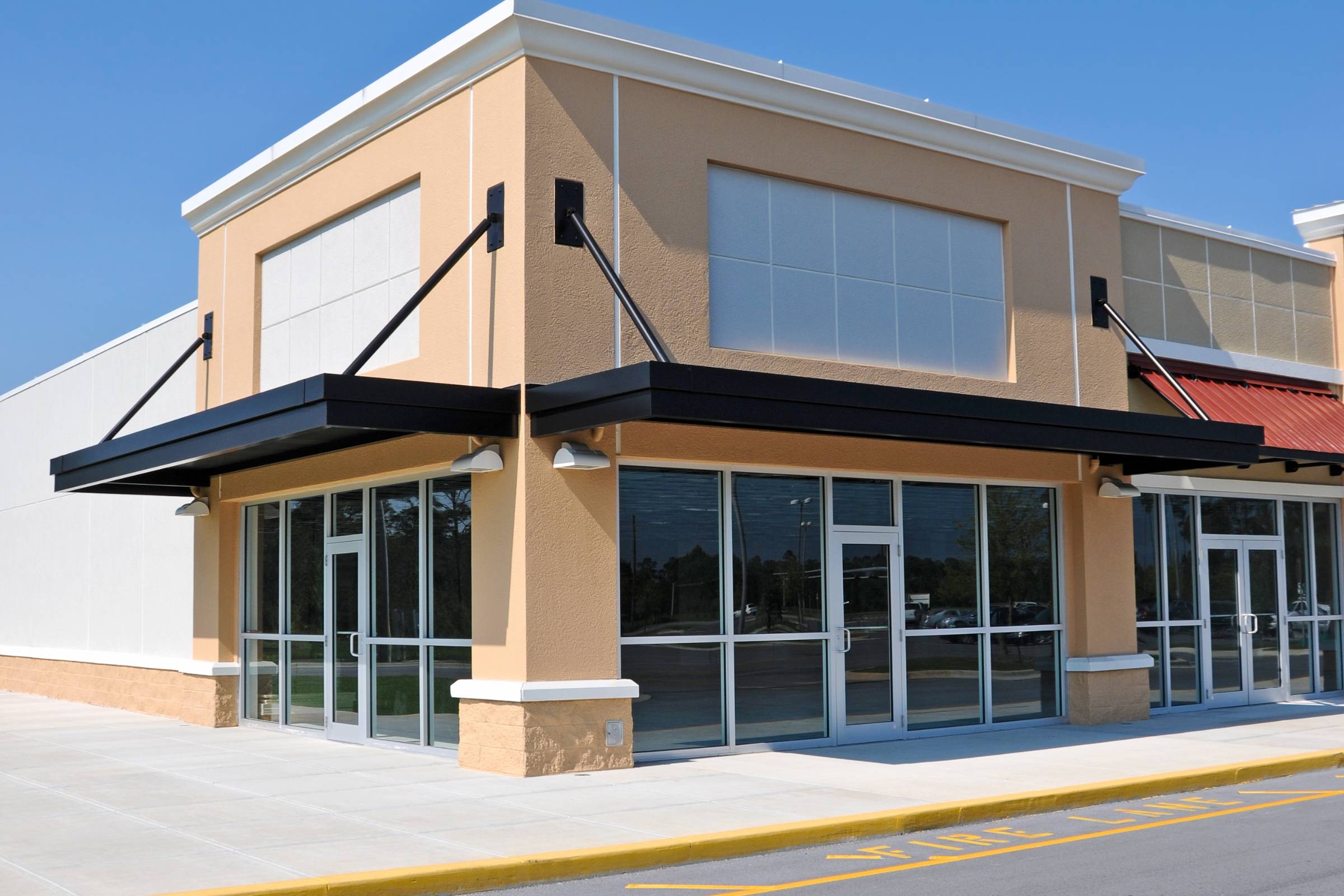 A vacant storefront with large windows and glass doors in a modern commercial building, featuring new commercial interior and exterior painting, located on a sunny day with a clear blue sky.
