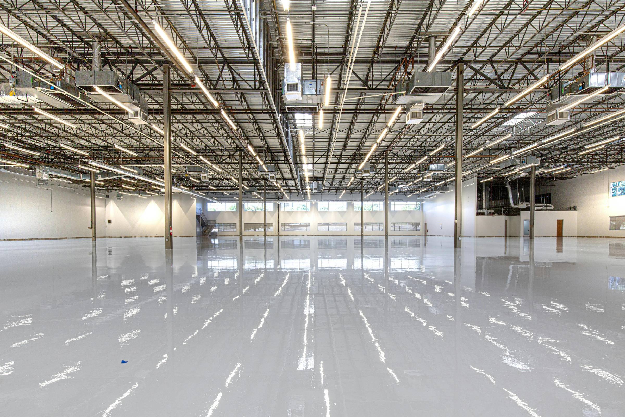 Large, empty industrial warehouse with a shiny, polished concrete floor, exposed ceiling beams, bright overhead lighting, and fresh new construction painting throughout.