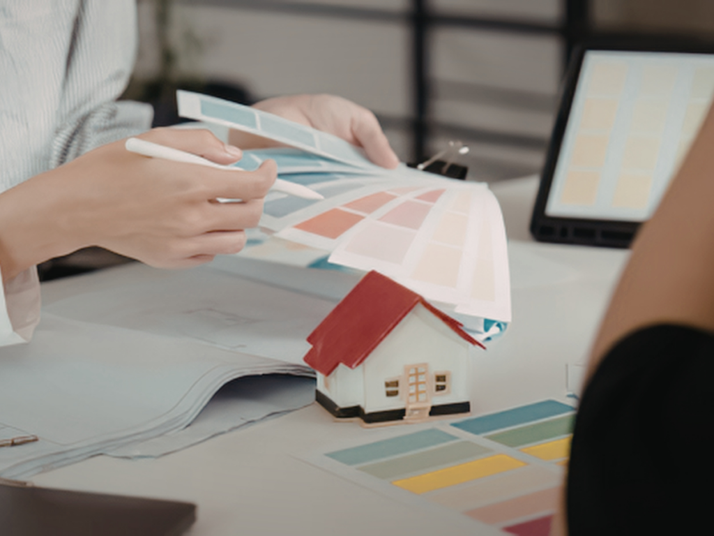 Person selecting paint swatches at a desk with a model house and an open folder, suggesting how to choose the perfect paint color for an interior design or home renovation planning session.
