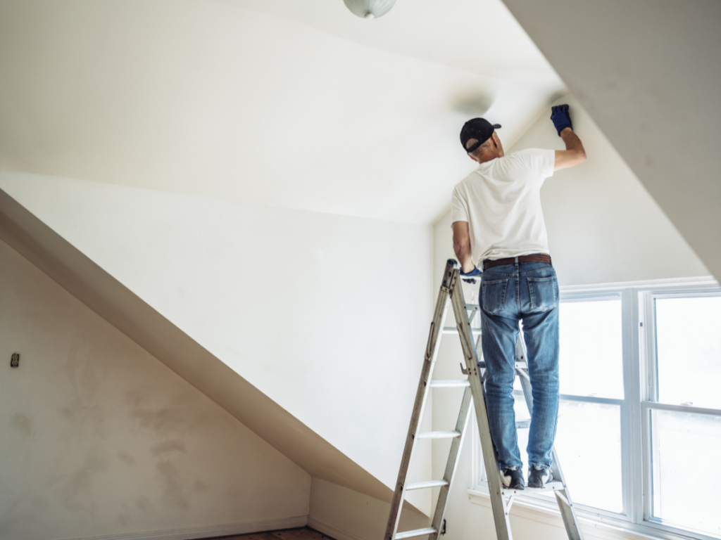 A painting contractor from Marietta GA stands on a ladder, painting the upper walls and ceiling of a white room with angled ceilings and a window.
