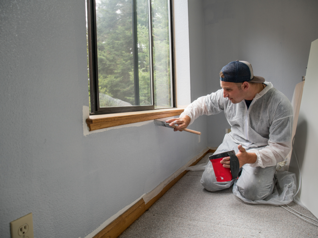 A painting contractor in Marietta GA, dressed in coveralls, kneels by a window, painting a light gray wall with a brush and holding a red paint tray.