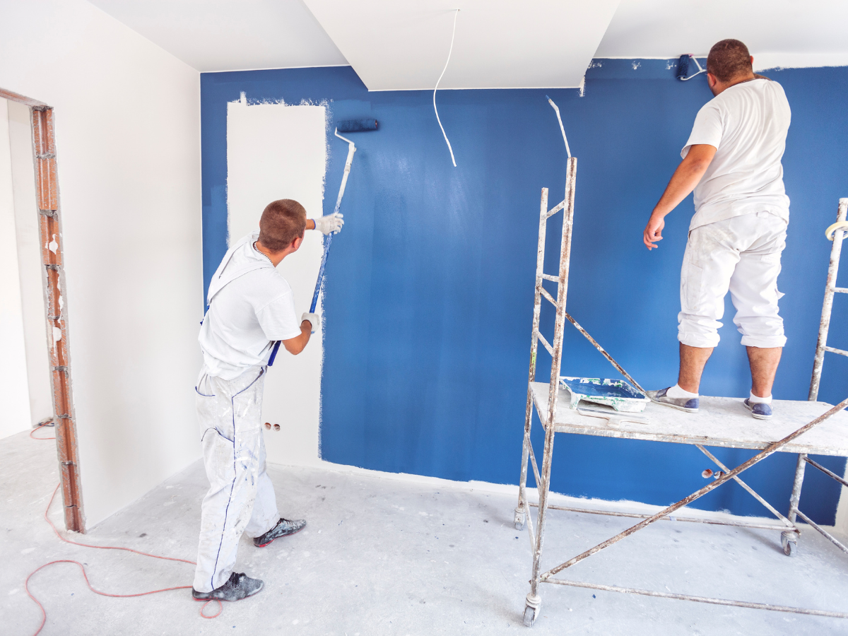 Two workers in white overalls from a painting contractor Marietta GA are painting an interior wall blue—one uses a roller on a ladder, while the other carefully paints near the corner on the floor.
