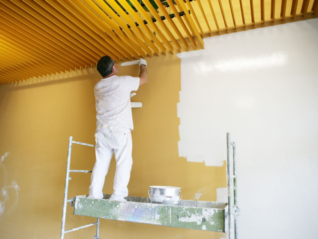 A person standing on scaffolding paints a yellow wall white with a roller brush, beneath a ceiling with wooden slats—showcasing the difference between professional painting vs DIY painting results.
