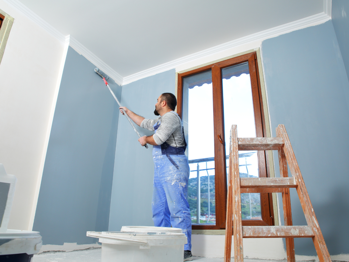 A person wearing blue overalls paints a light blue wall with a paint roller, highlighting the difference between professional painting vs DIY painting in a room with a ladder, window, and paint supplies on the floor.