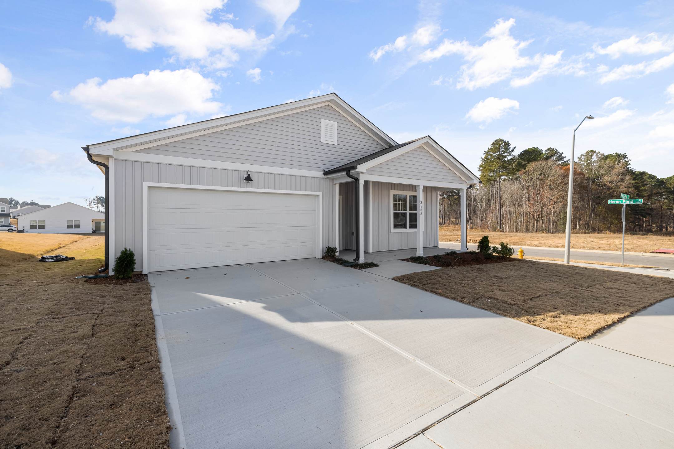 Single-story house with light gray siding, a white garage door, and a covered front entrance, featuring residential epoxy coatings on the concrete driveway. Situated on a corner lot with a small lawn.