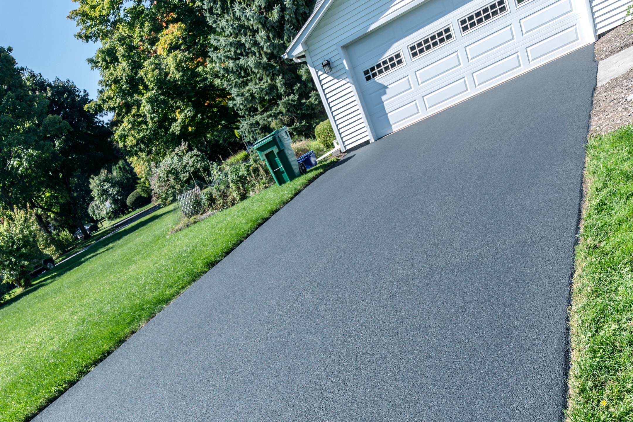 Freshly paved asphalt driveway leading to a white garage with three doors, enhanced with residential epoxy coatings, surrounded by green grass and trees on a sunny day.