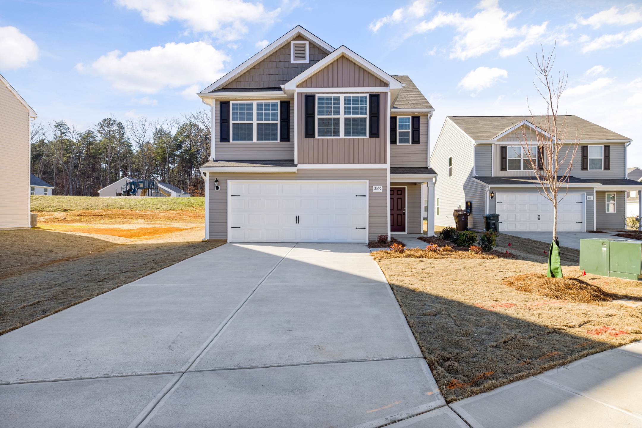 Two-story suburban house with beige siding, white garage door, and black shutters. Concrete driveway leads to the entrance, ideal for residential epoxy coatings. Neighboring houses and clear sky in the background.