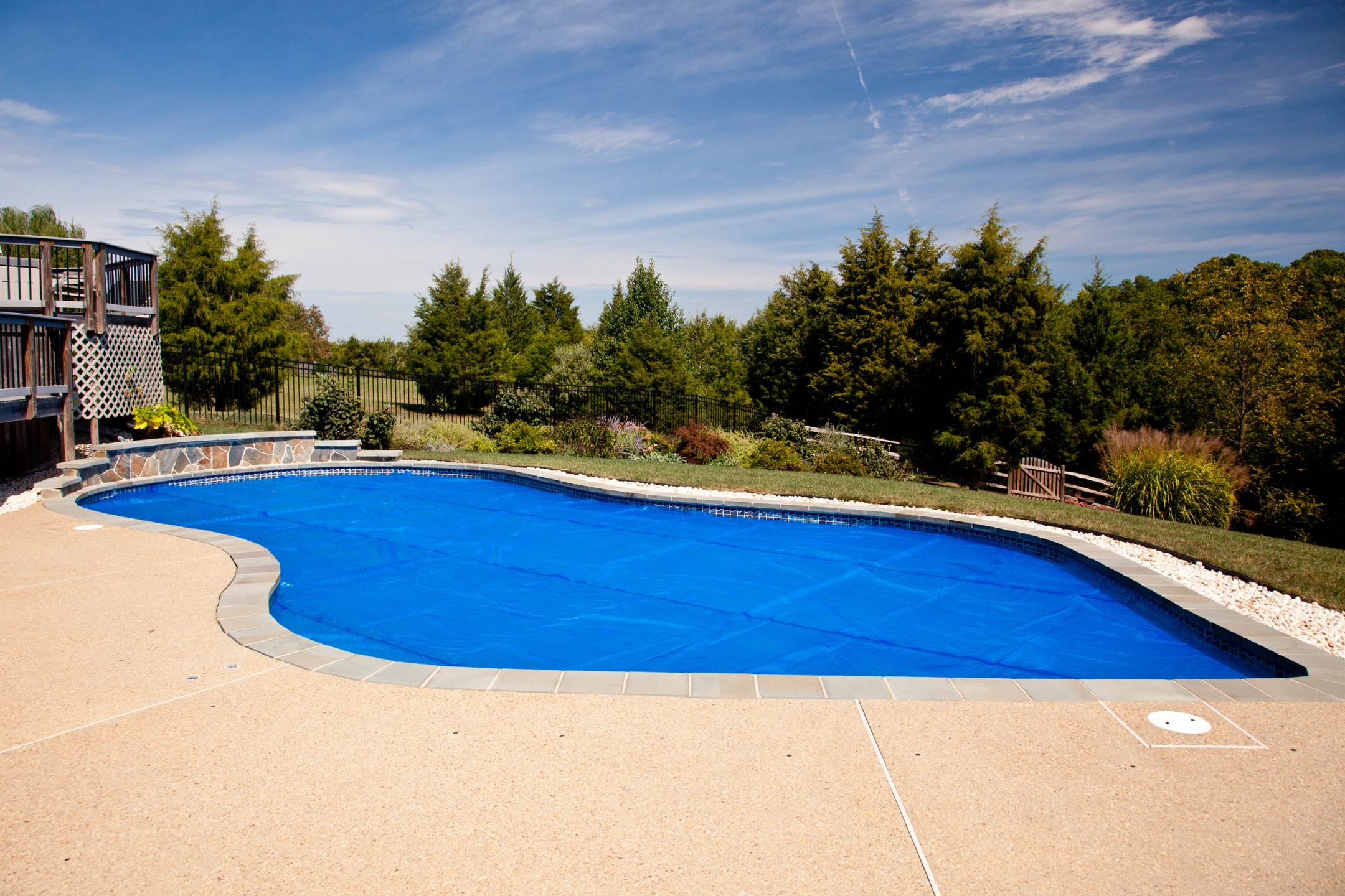 An outdoor swimming pool with a blue cover on, surrounded by a concrete deck finished with residential epoxy coatings and landscaping with trees in the background.