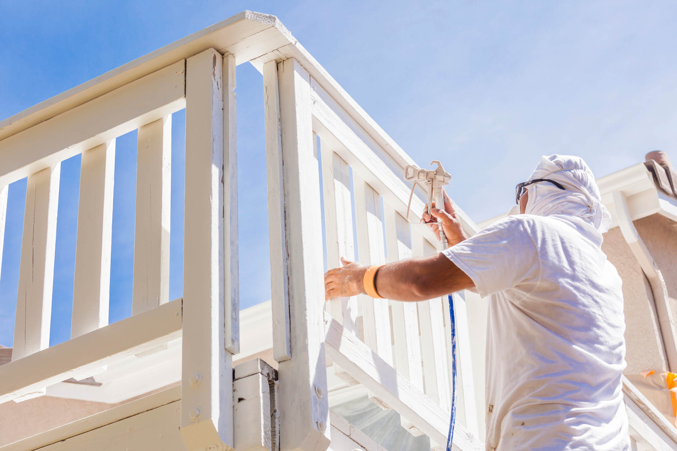 Wearing protective clothing, a person uses a paint sprayer to coat an outdoor wooden railing with white paint, showcasing precision in new construction painting on a sunny day.